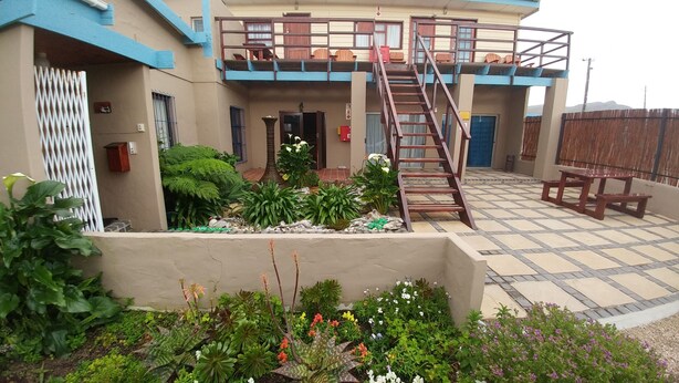 A two-story building with a balcony, a staircase, and a garden area with various plants and flowers. - Fisherhaven Traveller's Lodge - Hostel (Hermanus)