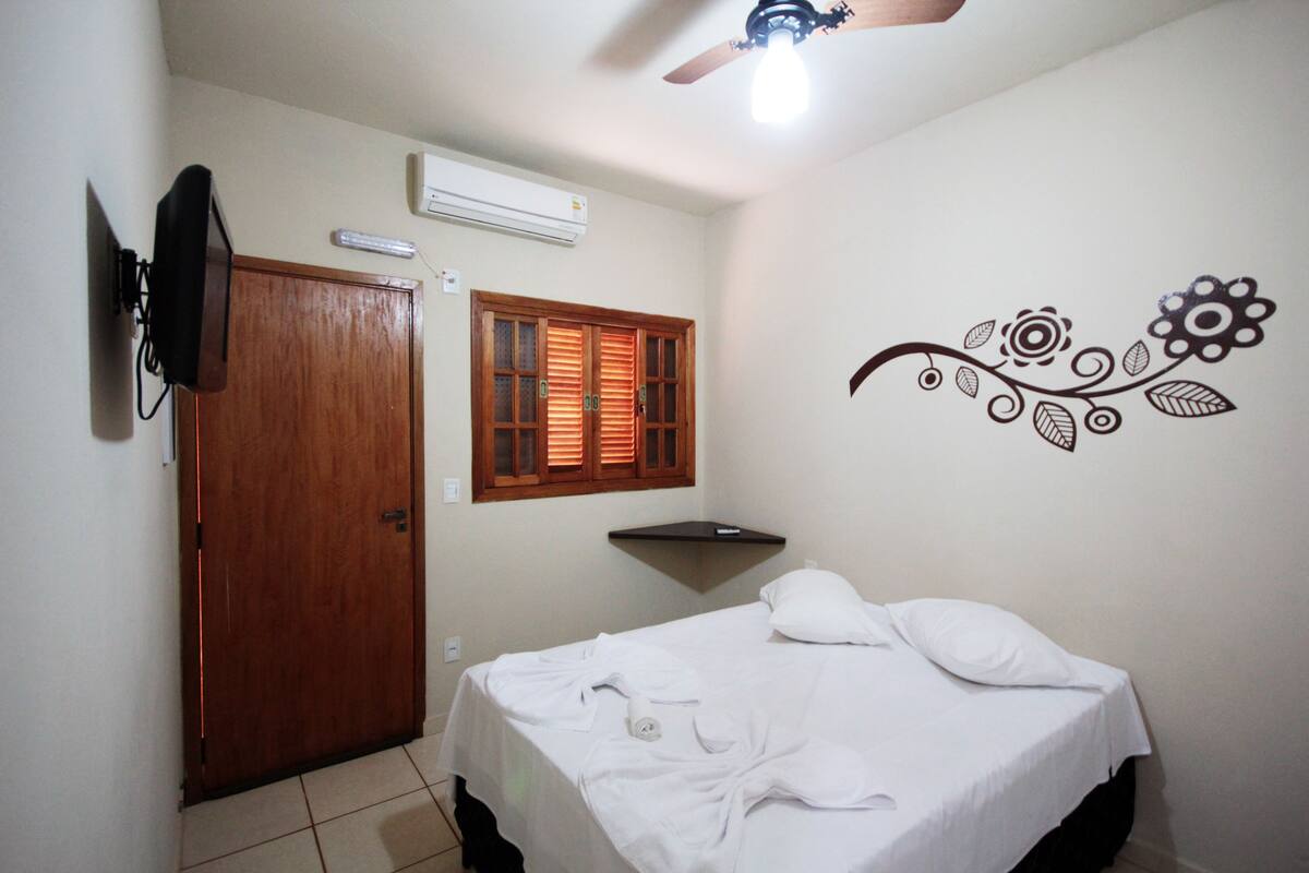 Une chambre à coucher avec un lit, une porte en bois, une fenêtre avec des stores, un téléviseur fixé au mur et un appareil de climatisation.