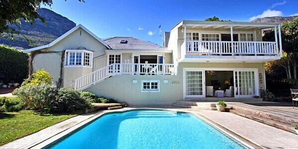 A two-story house with a swimming pool, a balcony, and a mountain backdrop.