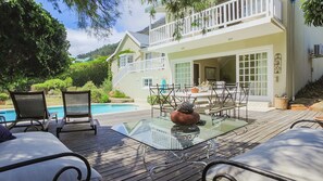 A poolside area with a glass table, chairs, and a view of a two-story house with a balcony.