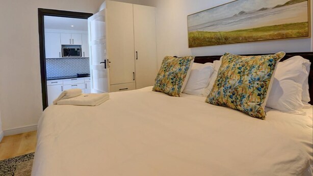 A bedroom with a large bed, two pillows, a painting on the wall, and a view into the kitchen. - Cloverleigh Guest House (Wilderness)