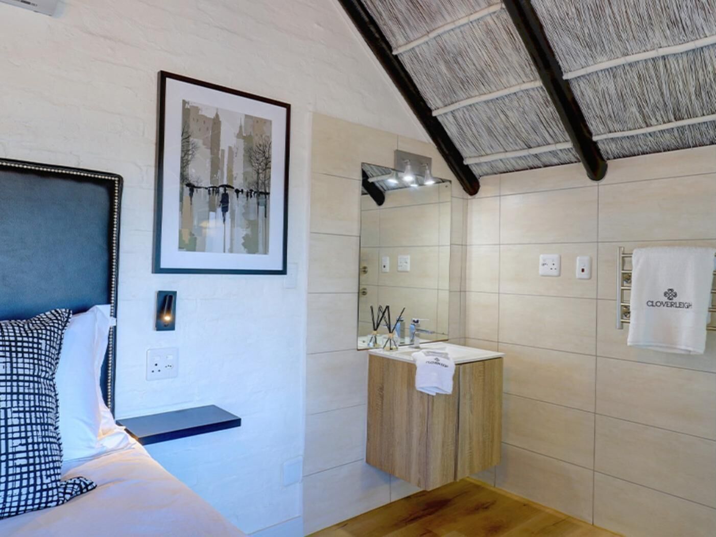 Une chambre à coucher comprenant un lit, une table de chevet, un tableau encadré, ainsi qu’une salle de bain avec un lavabo, un miroir, un porte-serviettes et une douche.