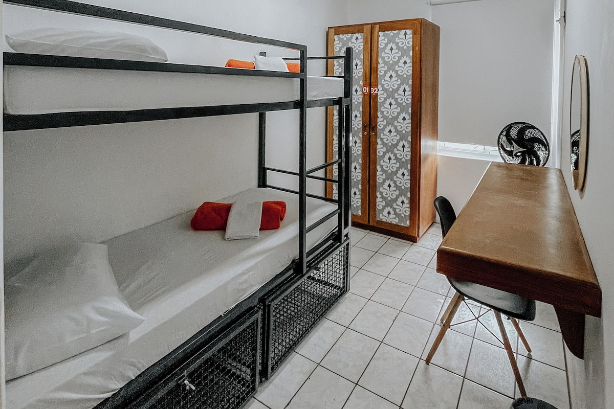 A compact room with bunk beds, a wooden table, a chair, a wardrobe, and a fan.