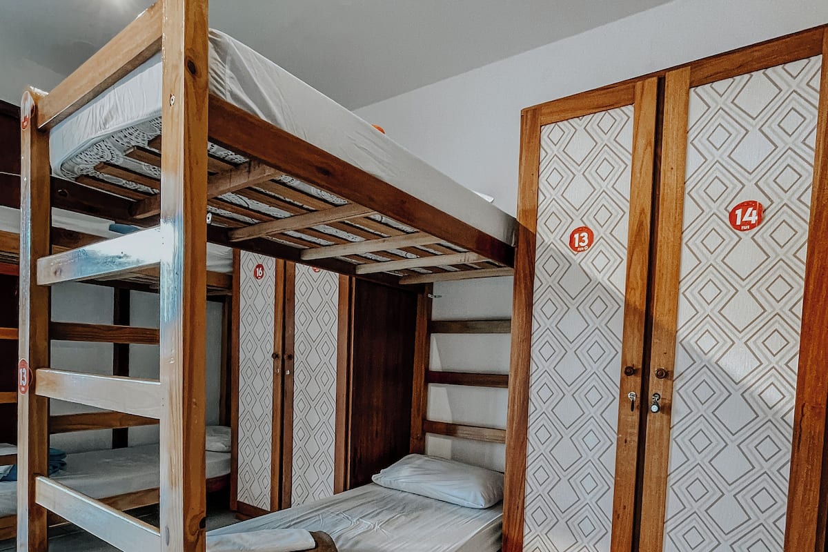 A bunk bed with a wooden frame and a lower bunk with a mattress. There is a wardrobe with a geometric pattern and a door marked with the number 14.
