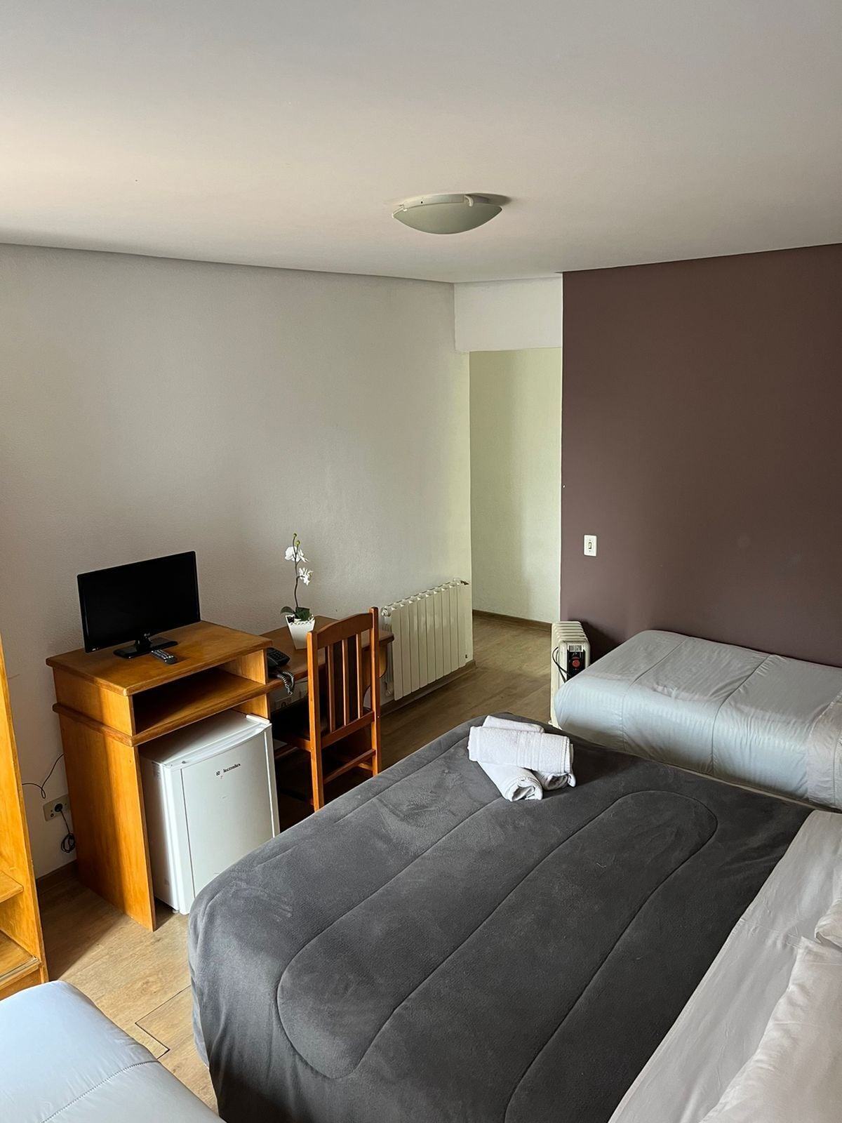 A hotel room with a bed, a desk with a computer, a small refrigerator, and a radiator.