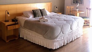 A bedroom with a wooden ceiling, a large bed with a white bedspread, a nightstand with a lamp, and a chair. - Pousada Condado Brasileiro (Campos do Jordão)