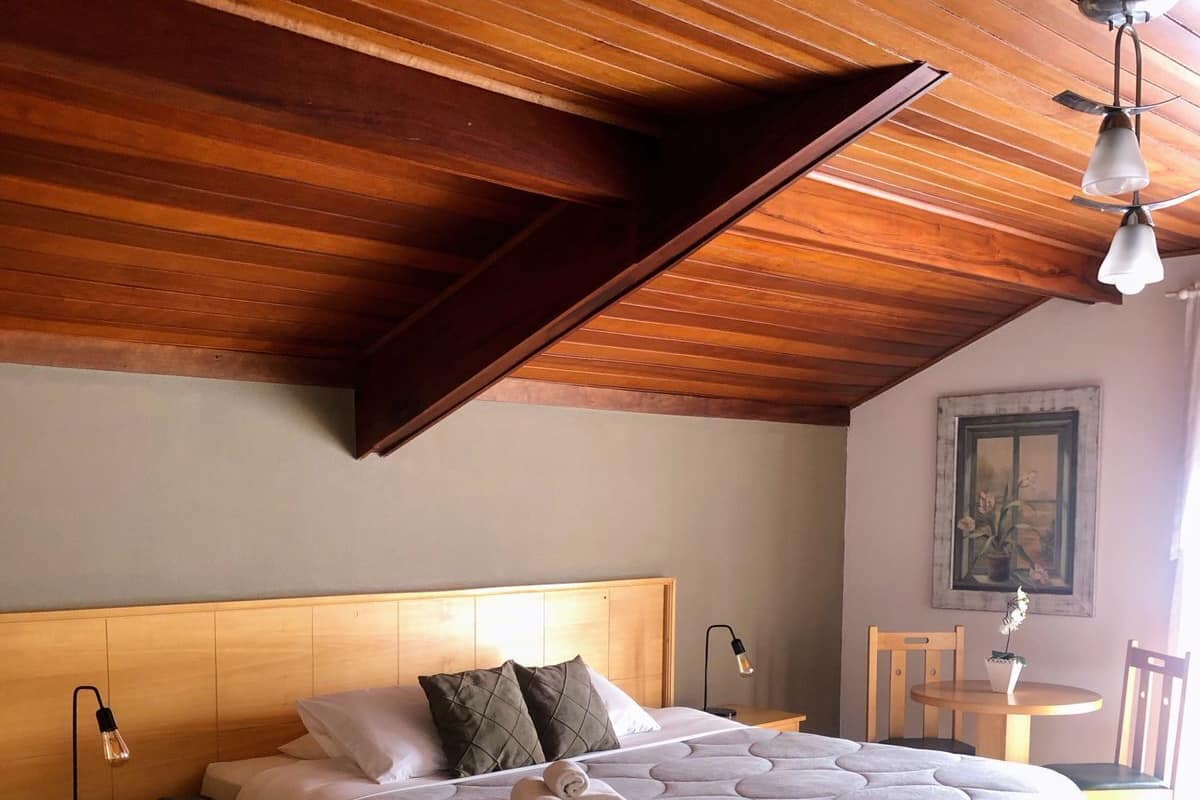 Un dormitorio con techo de madera, una cama grande con cubrecama blanco, una mesita de noche con lámpara y una silla.