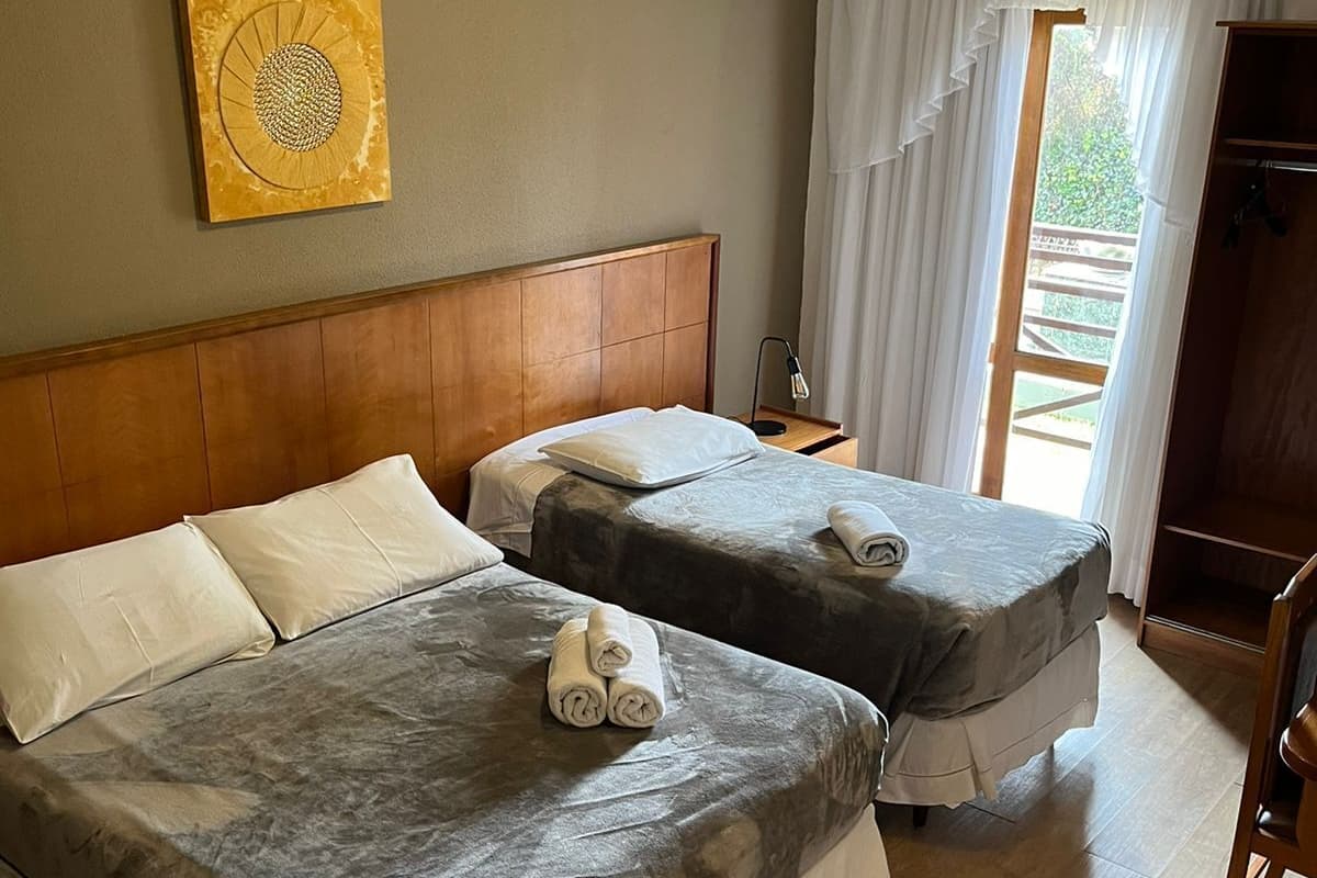 Una habitación de hotel con dos camas, cabecero de madera, mesita de noche con lámpara, un cuadro en la pared y una ventana con cortinas.