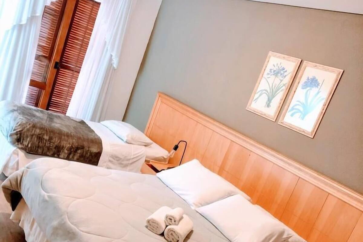Una cama doble con una colcha color beige, dos almohadas y toallas enrolladas. Hay una mesita de noche de madera con una lámpara, un cabecero de madera y dos láminas botánicas enmarcadas en la pared.