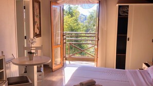 A bedroom with a bed, a bedside table, a chair, a small table with a lamp, and a balcony with a view of trees and mountains. - Pousada Condado Brasileiro (Campos do Jordão)