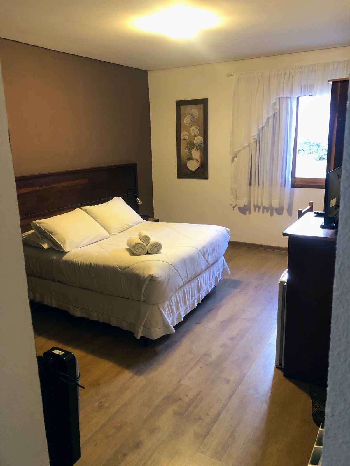 Um quarto com cama, escrivaninha, cadeira, janela com cortinas e um quadro na parede.