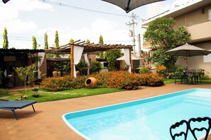 A swimming pool with a heart-shaped decoration, surrounded by a garden with various plants and a pergola structure.