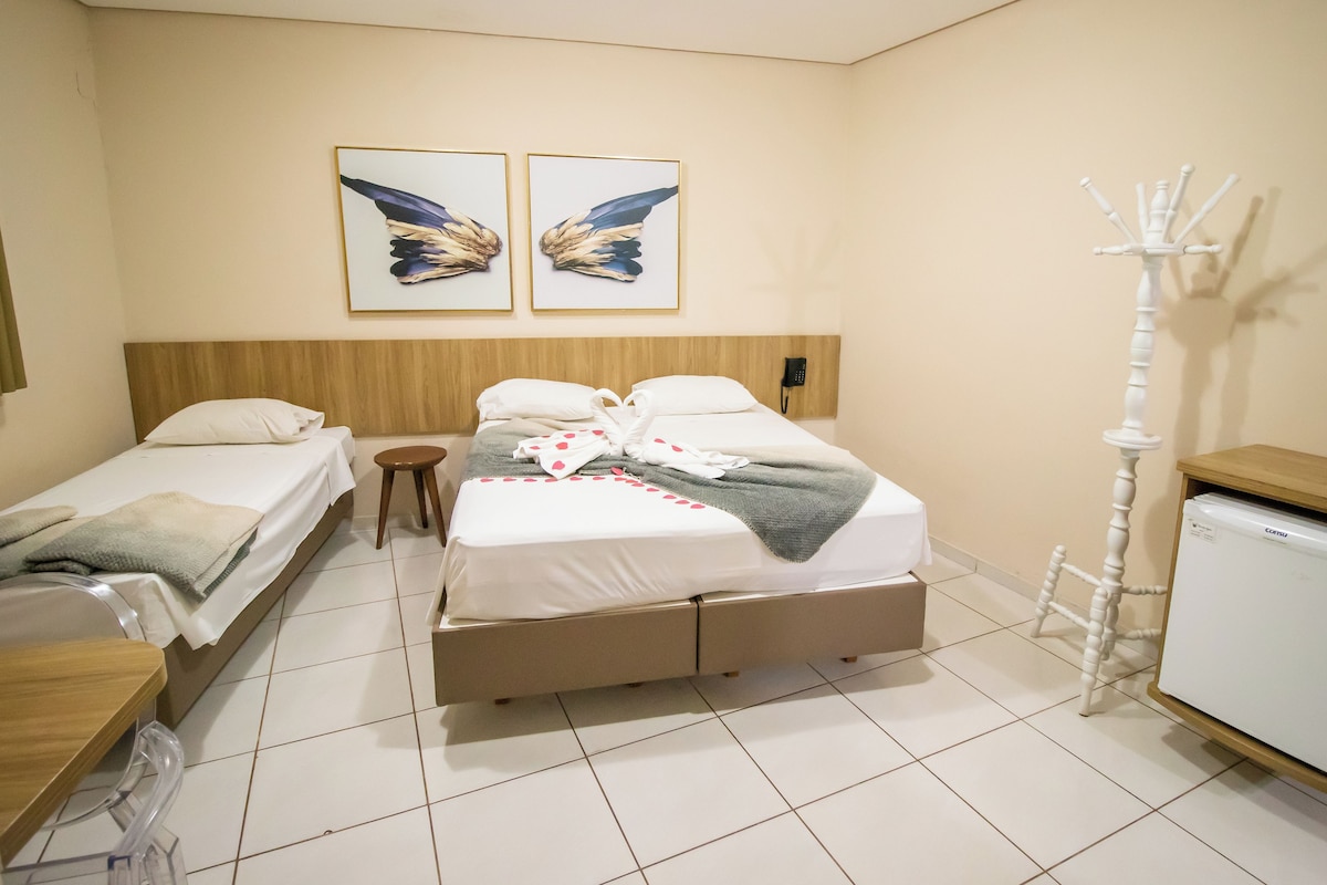 Quarto de hotel com uma cama de casal, uma cama simples, cabeceira de madeira, mesa de cabeceira, um banquinho pequeno, um cabideiro e uma geladeira.