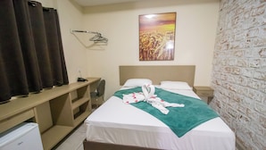 A hotel room with a bed, a desk, a chair, a small fridge, a wall-mounted towel rack, and a picture of a field.