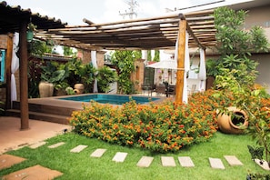 Quintal com piscina, cercado por um jardim com flores coloridas e uma pérgola.