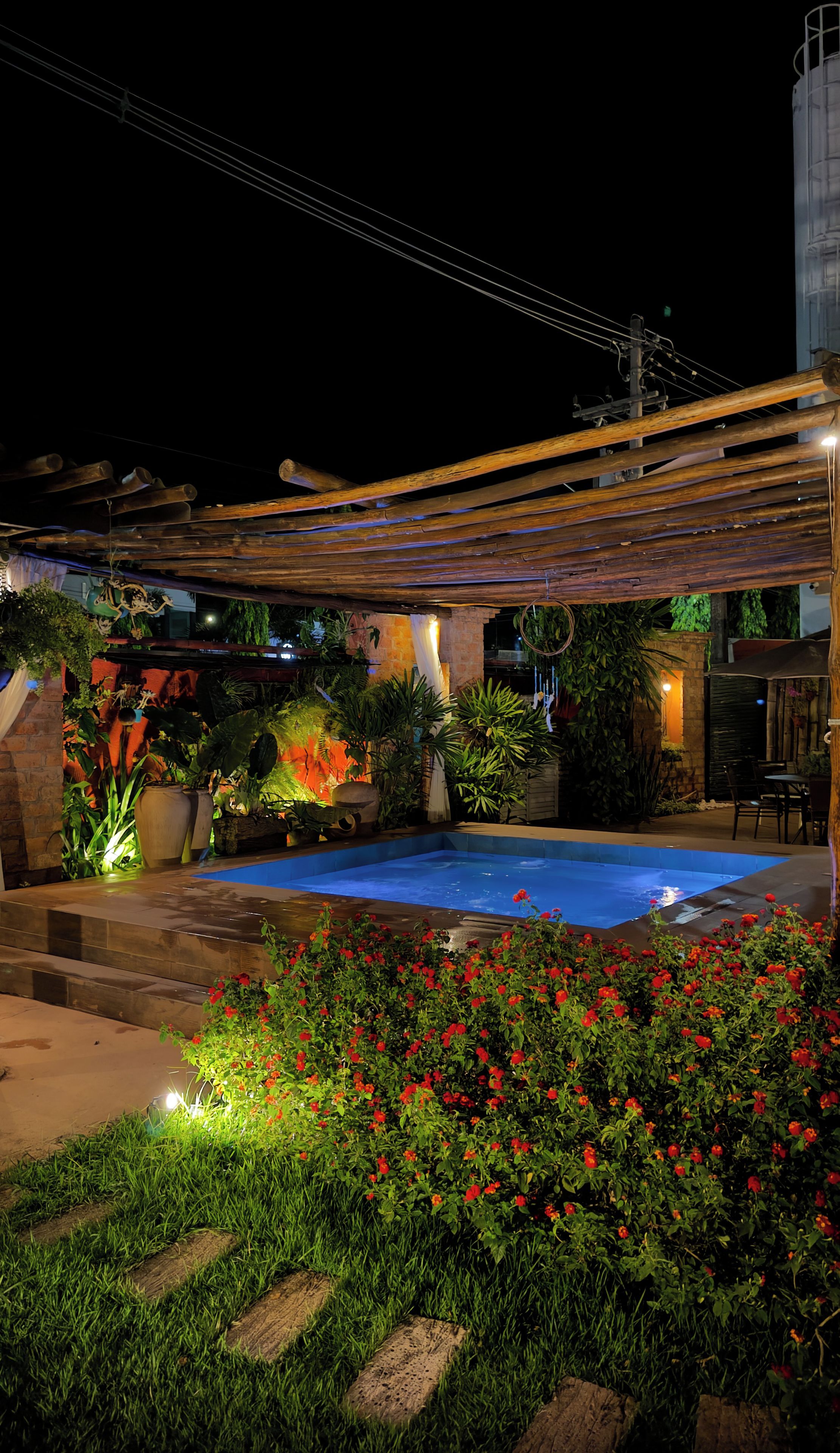 Zona de piscina exterior en la noche, con un pérgola, escalones que bajan a la piscina y diversas plantas y flores.