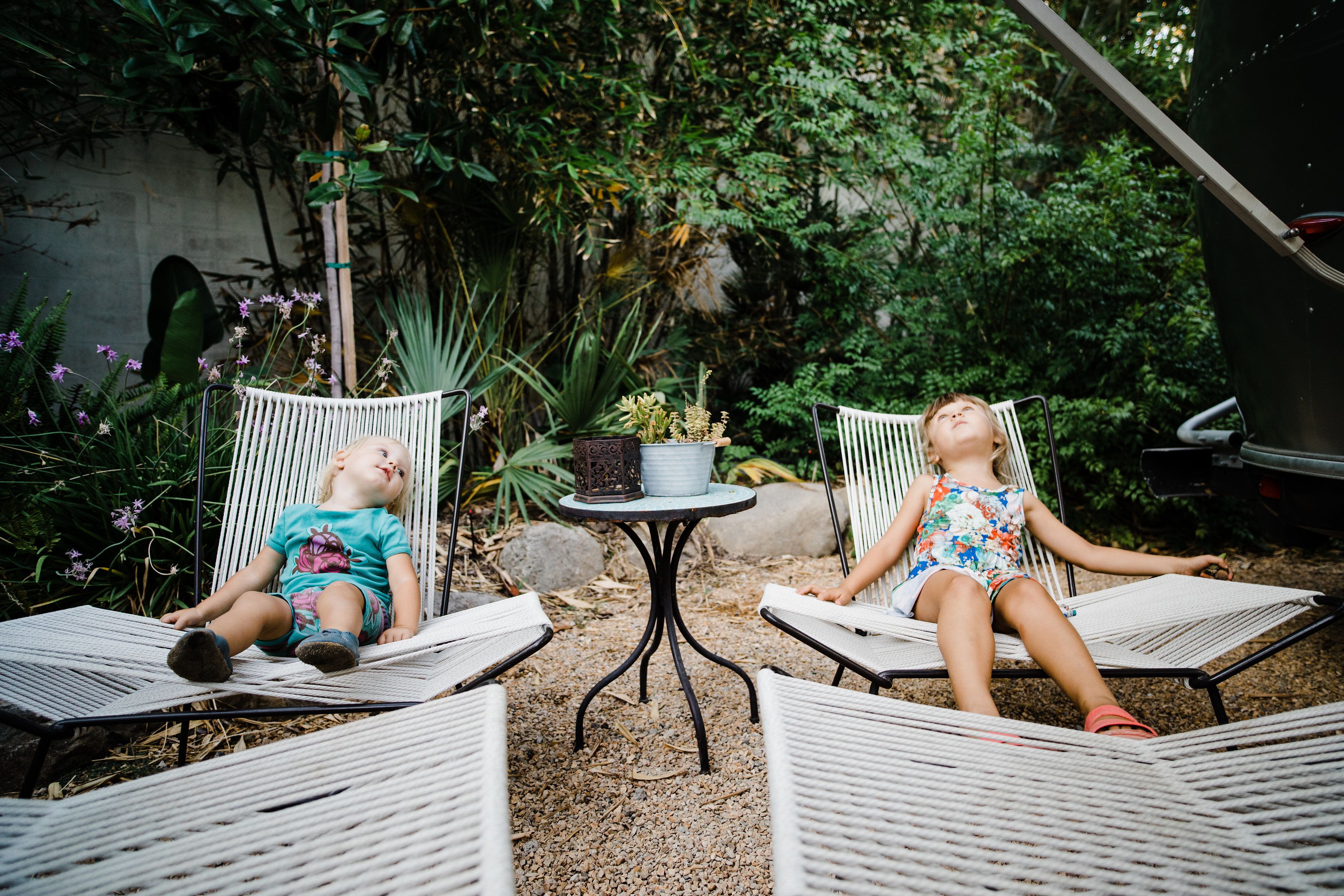 Dos niños sentados en sillas de mimbre en un jardín, con una pequeña mesa y plantas en macetas entre ellos.