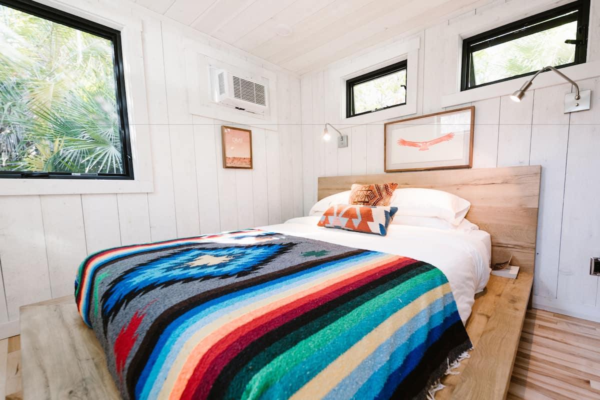 A bedroom with a bed, a colorful blanket, a wooden headboard, and framed artwork on the wall.