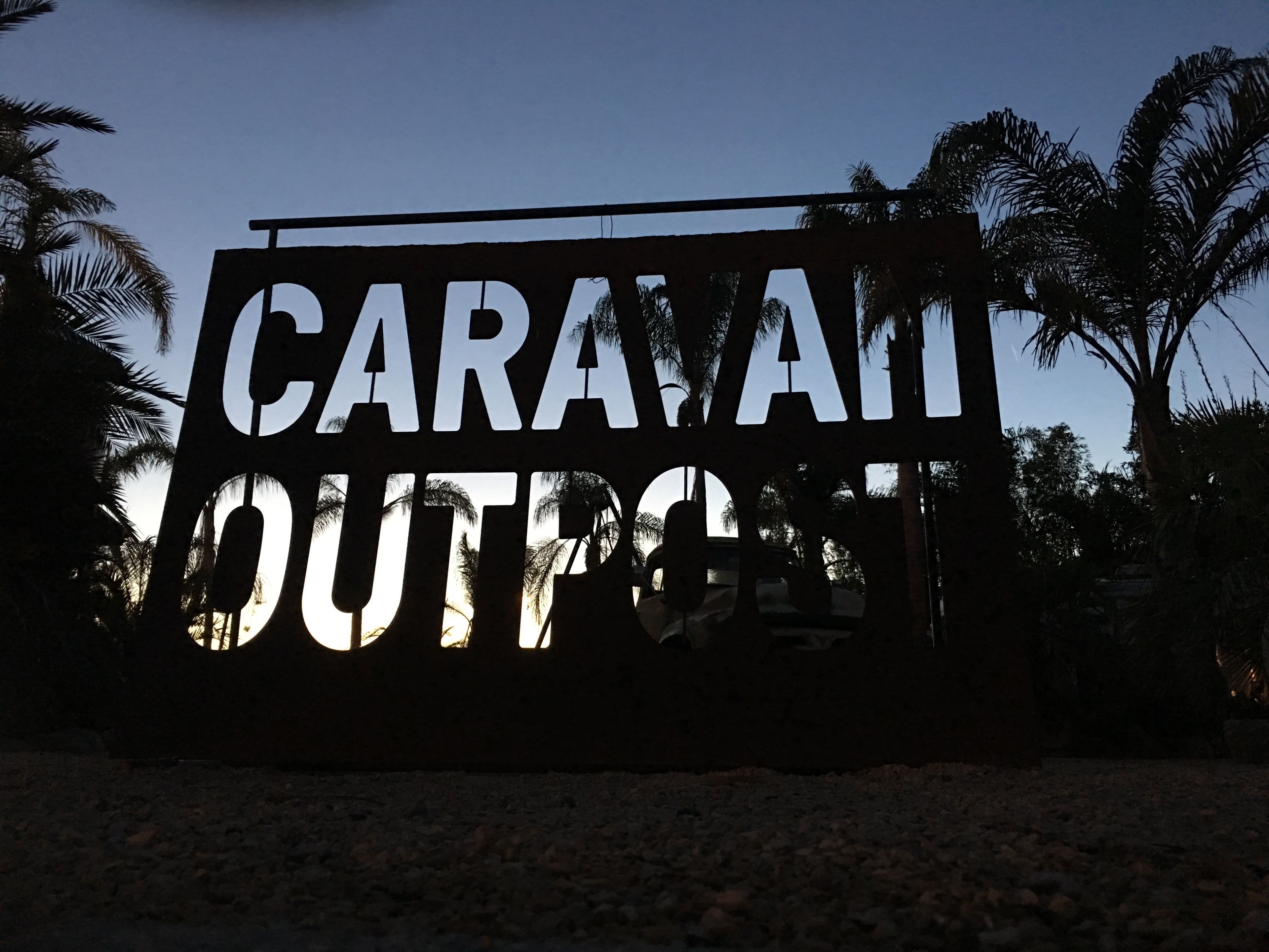 A sign with the text "CARAVAN OUTPOST" against a backdrop of palm trees and a clear sky.