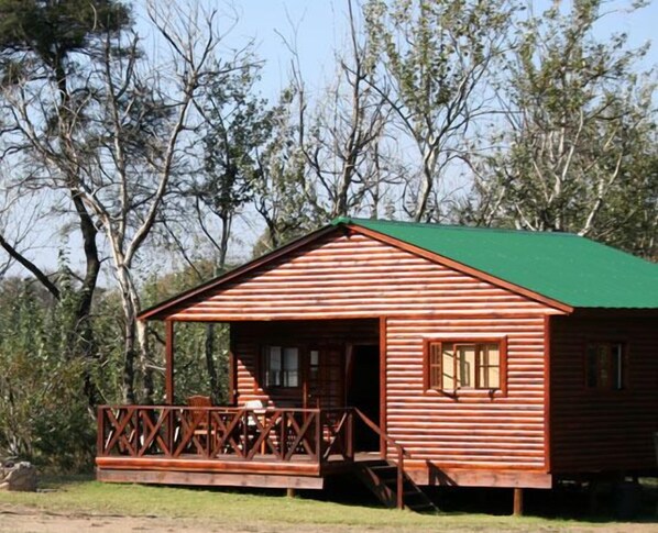 A wooden cabin with a green roof, a porch with a railing, and a staircase leading to the entrance. - Intaba Thulile (Maanhaarand)