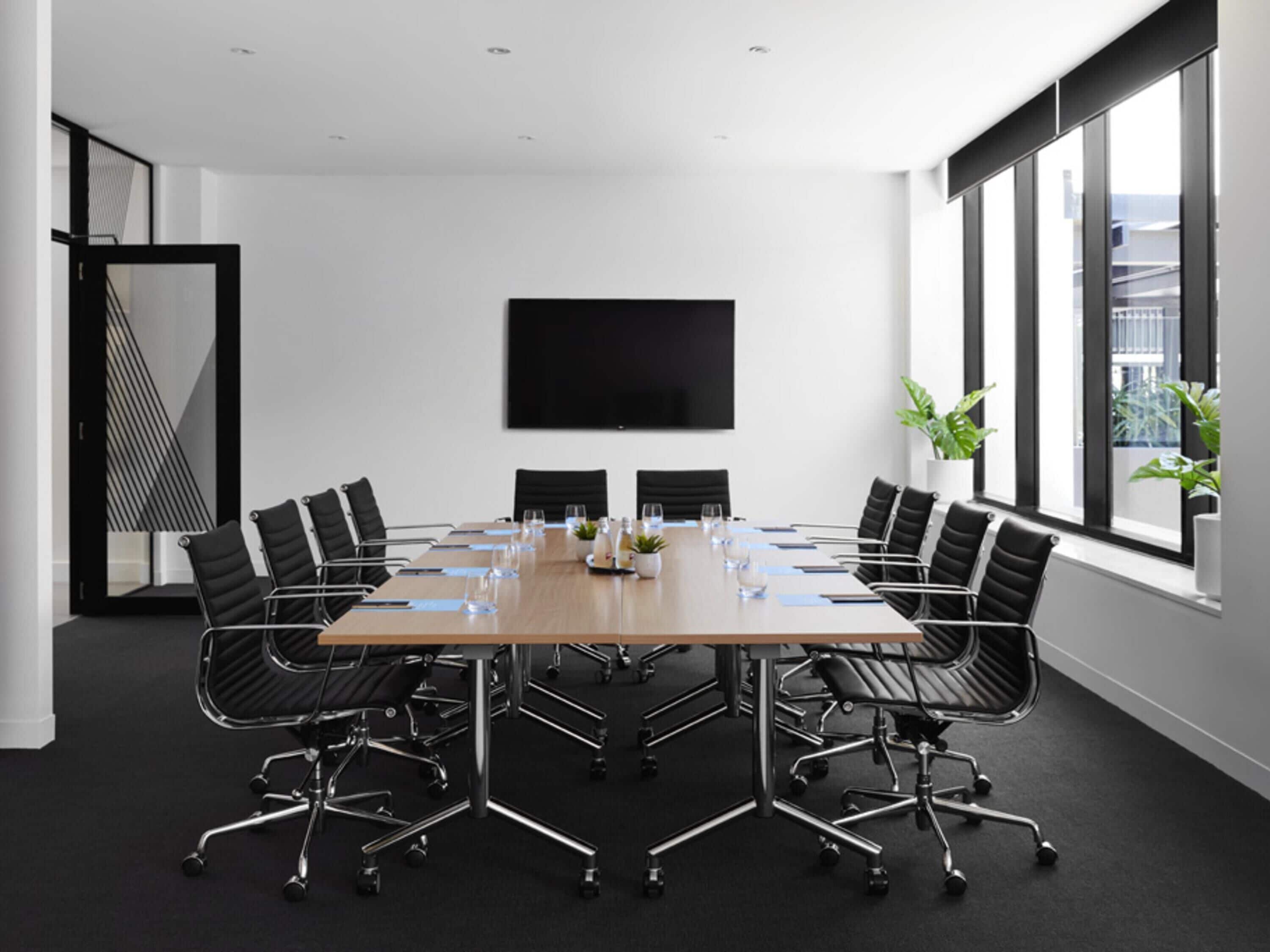 A conference room with a long table, black chairs, a flat-screen TV, and large windows.