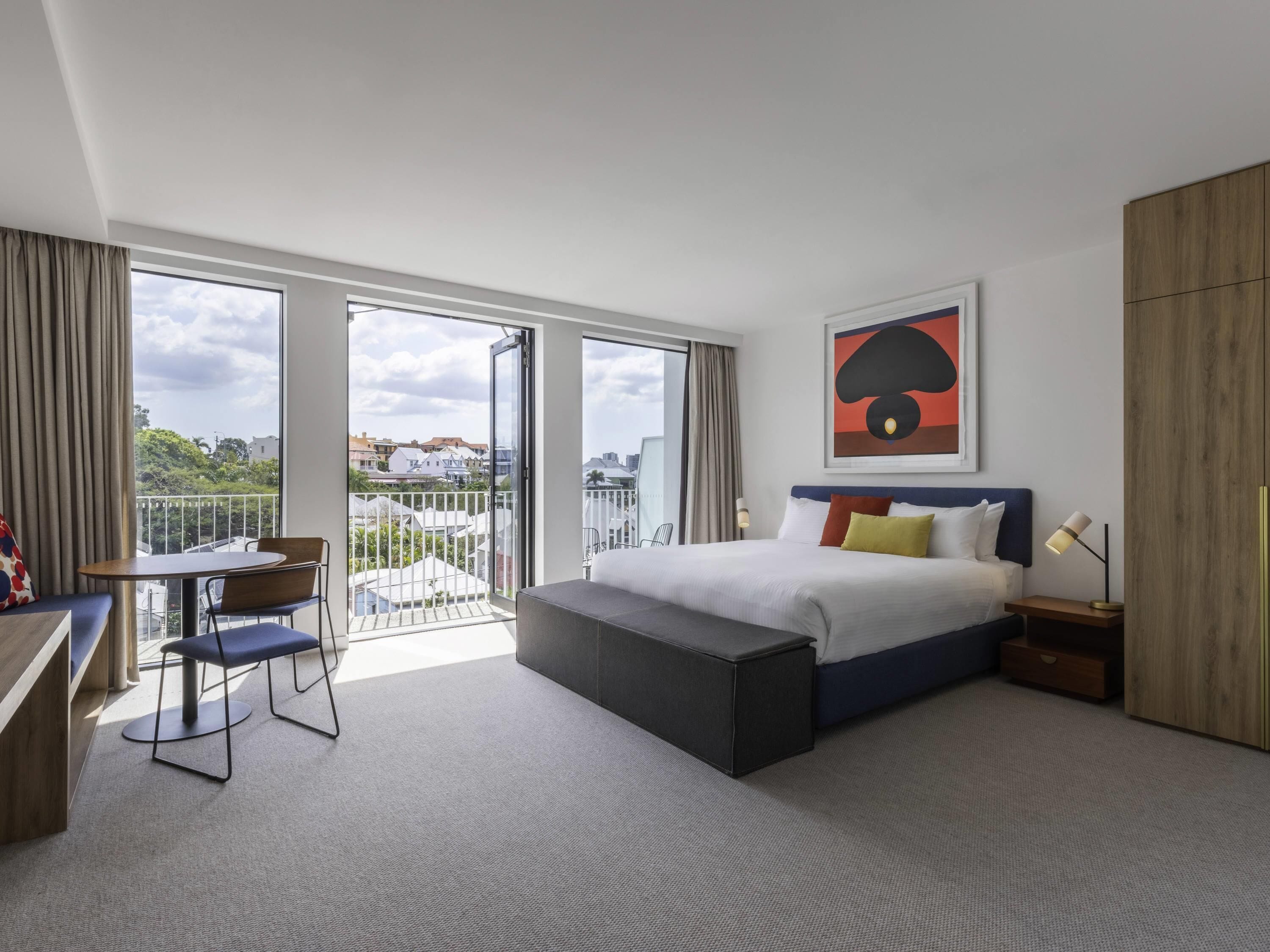 A modern hotel room with a large bed, a desk with a chair, a balcony with a view, and a painting on the wall.