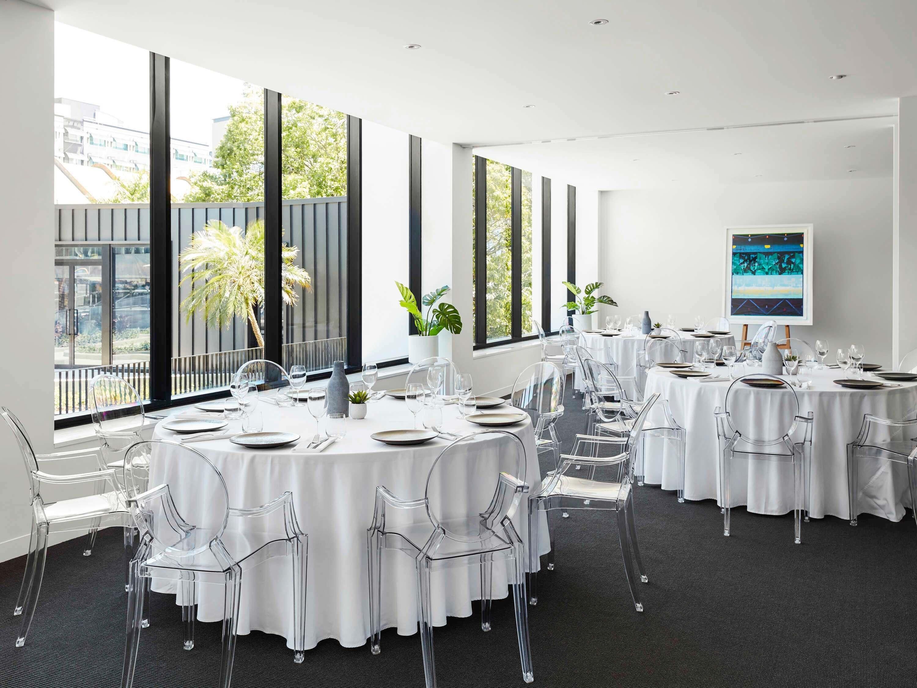 Un salón de conferencias con mesas redondas, sillas transparentes y amplias ventanas que dan vistas a edificios y árboles.