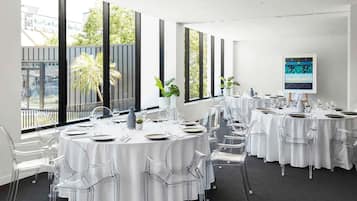 Un salón de conferencias con mesas redondas, sillas transparentes y amplias ventanas que dan vistas a edificios y árboles.