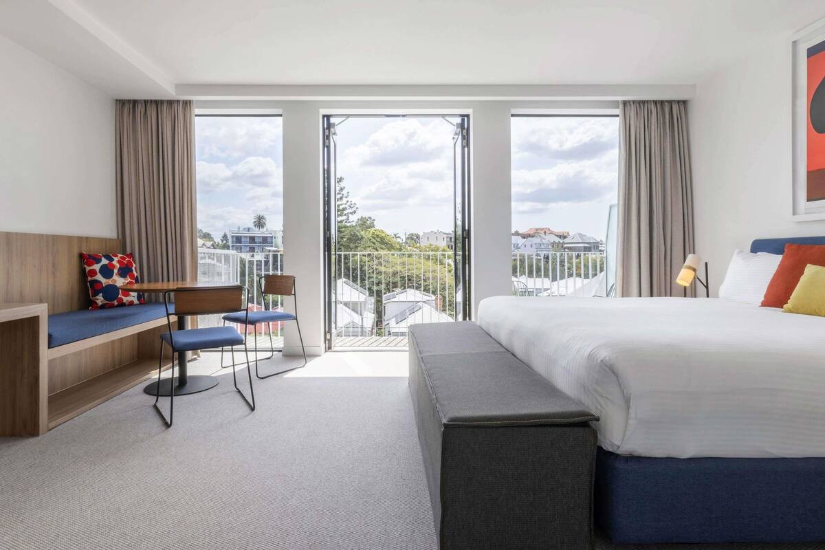 Habitación de hotel con una cama, una zona de estar con una mesa y sillas, un banco y ventanas amplias que ofrecen vistas a edificios y árboles.