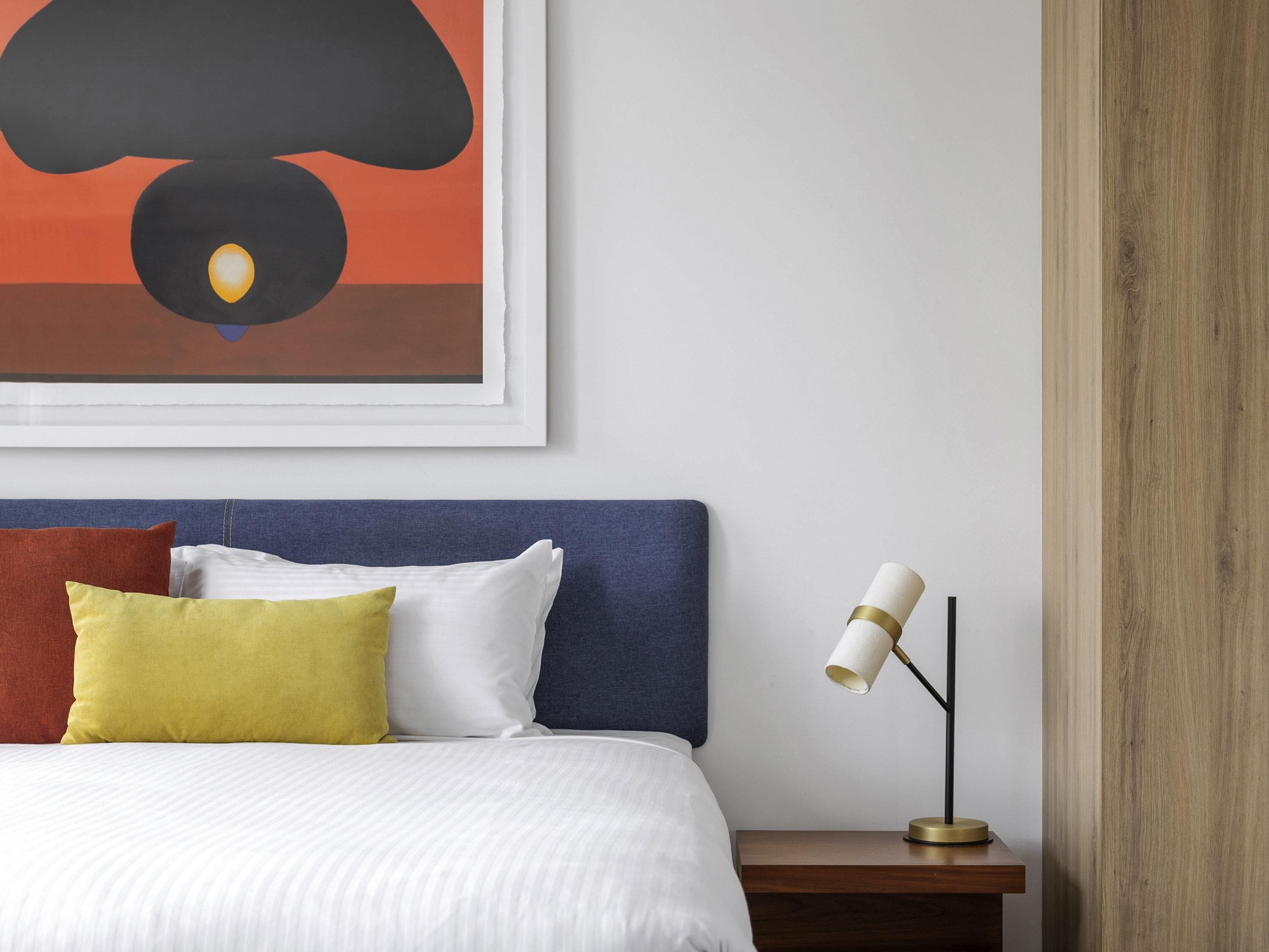 A neatly made bed with white and yellow pillows, a blue headboard, and a bedside table with a lamp.
