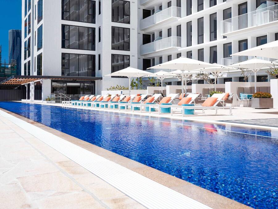 Une piscine entourée de chaises longues et de parasols, le tout bordé d’un bâtiment moderne doté de balcons.