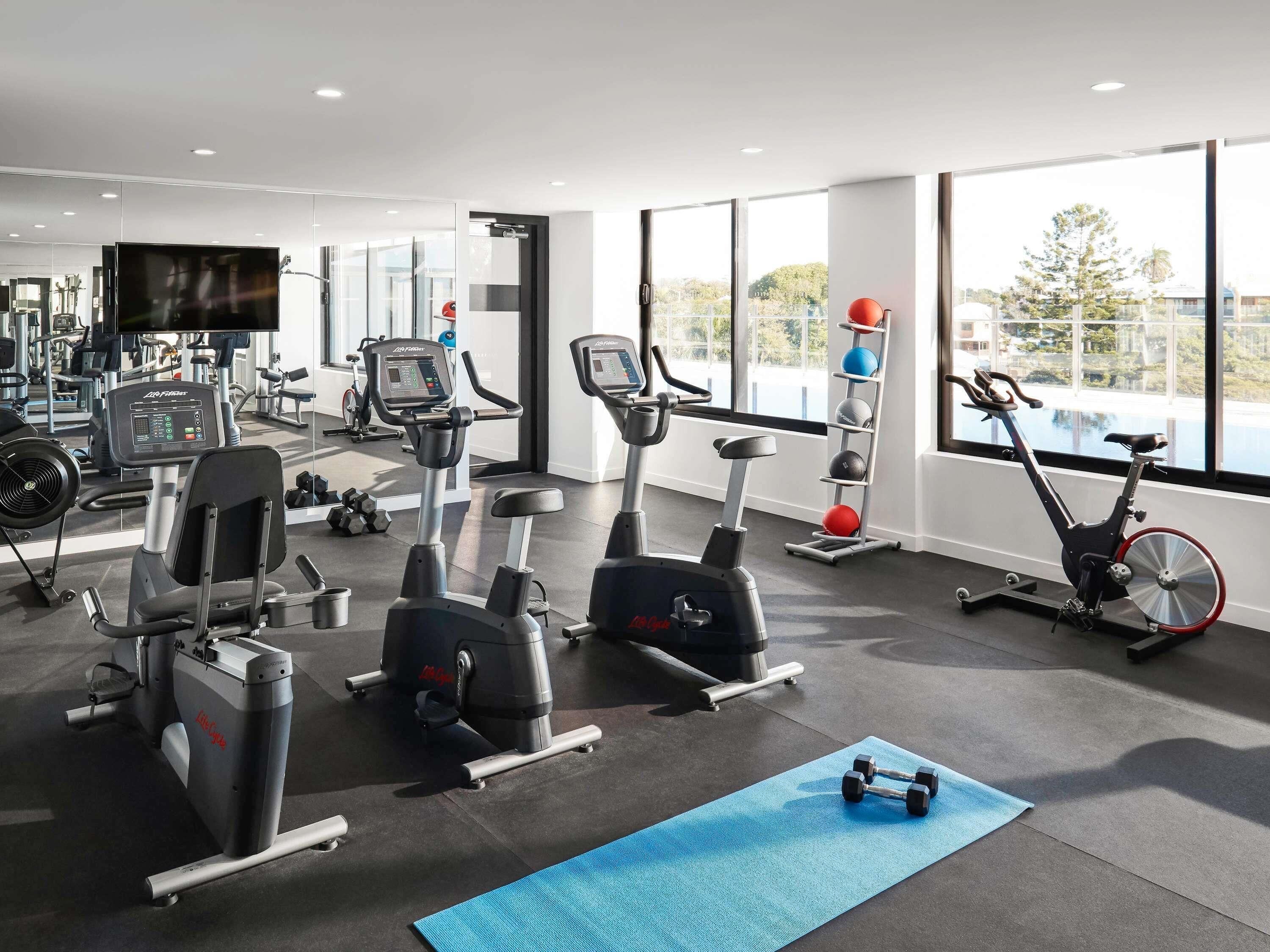 Un gimnasio con máquinas de cardio, un televisor en la pared y una ventana que ofrece vistas a árboles y edificios.