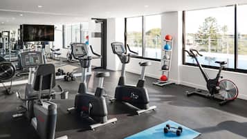 Un gimnasio con máquinas de cardio, un televisor en la pared y una ventana que ofrece vistas a árboles y edificios.