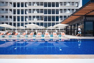 A swimming pool with lounge chairs and umbrellas, a modern building with balconies, and a person walking by.