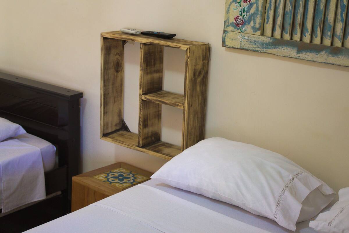 Une chambre avec une table de chevet en bois, un lit avec une literie blanche et une étagère fixée au mur, sur laquelle se trouve une télécommande.