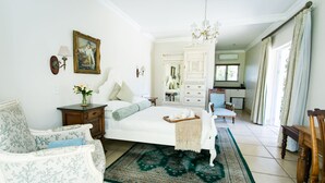 A spacious bedroom with a large bed, a sitting area with a chair, a dresser, and a view of the kitchen.