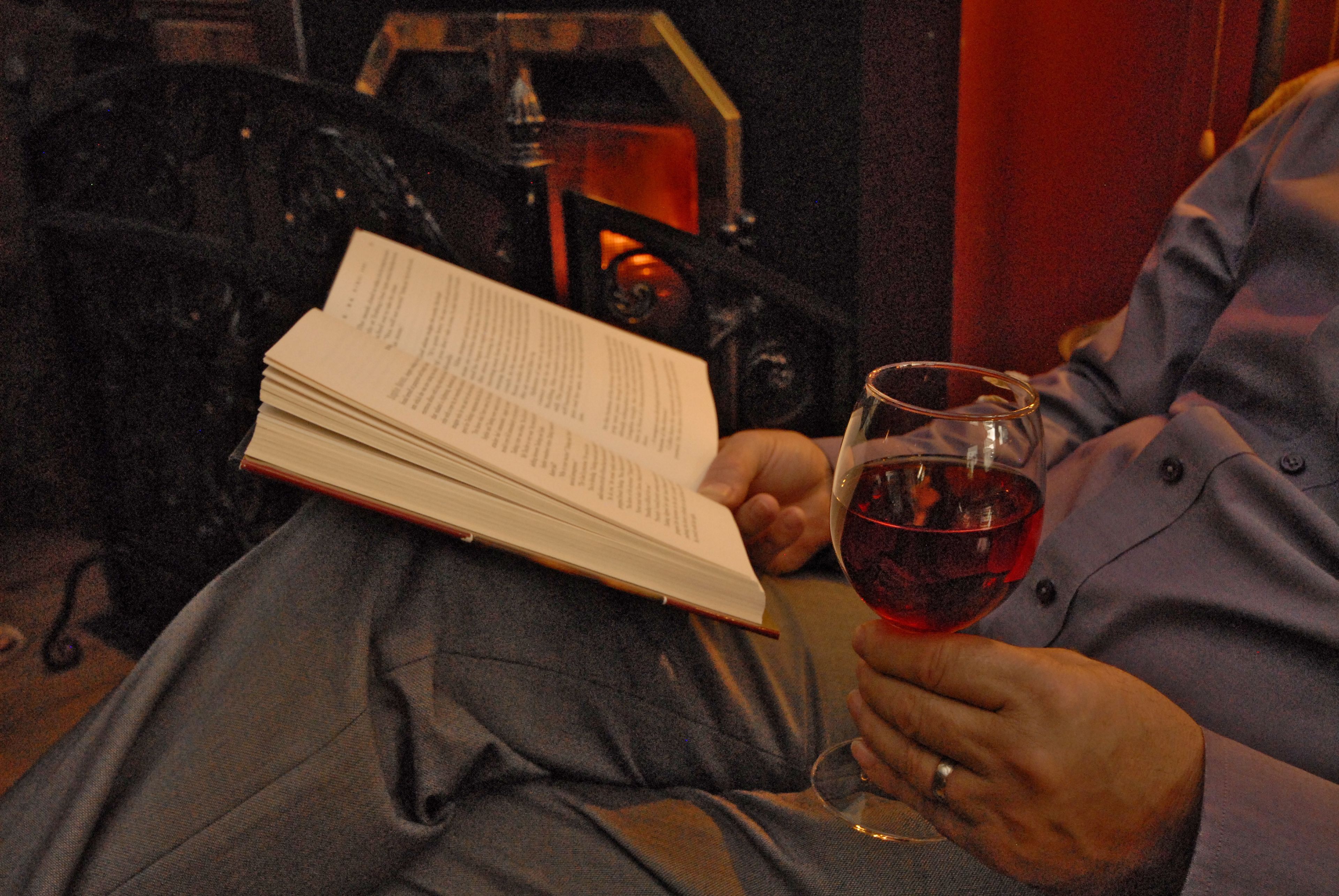 A person reading a book and holding a glass of red wine.