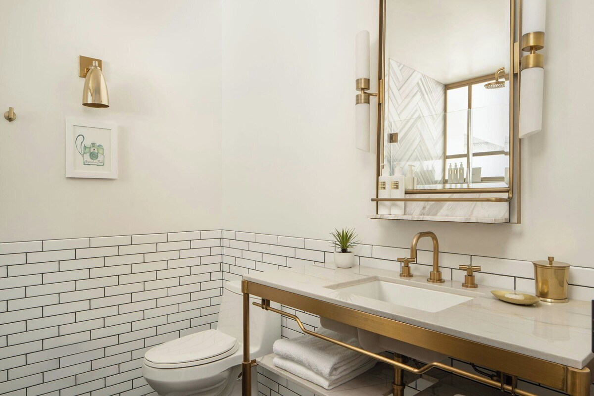 Un baño moderno con una paleta de colores blanco y dorado, que incluye un espejo grande, un lavamanos y un inodoro.