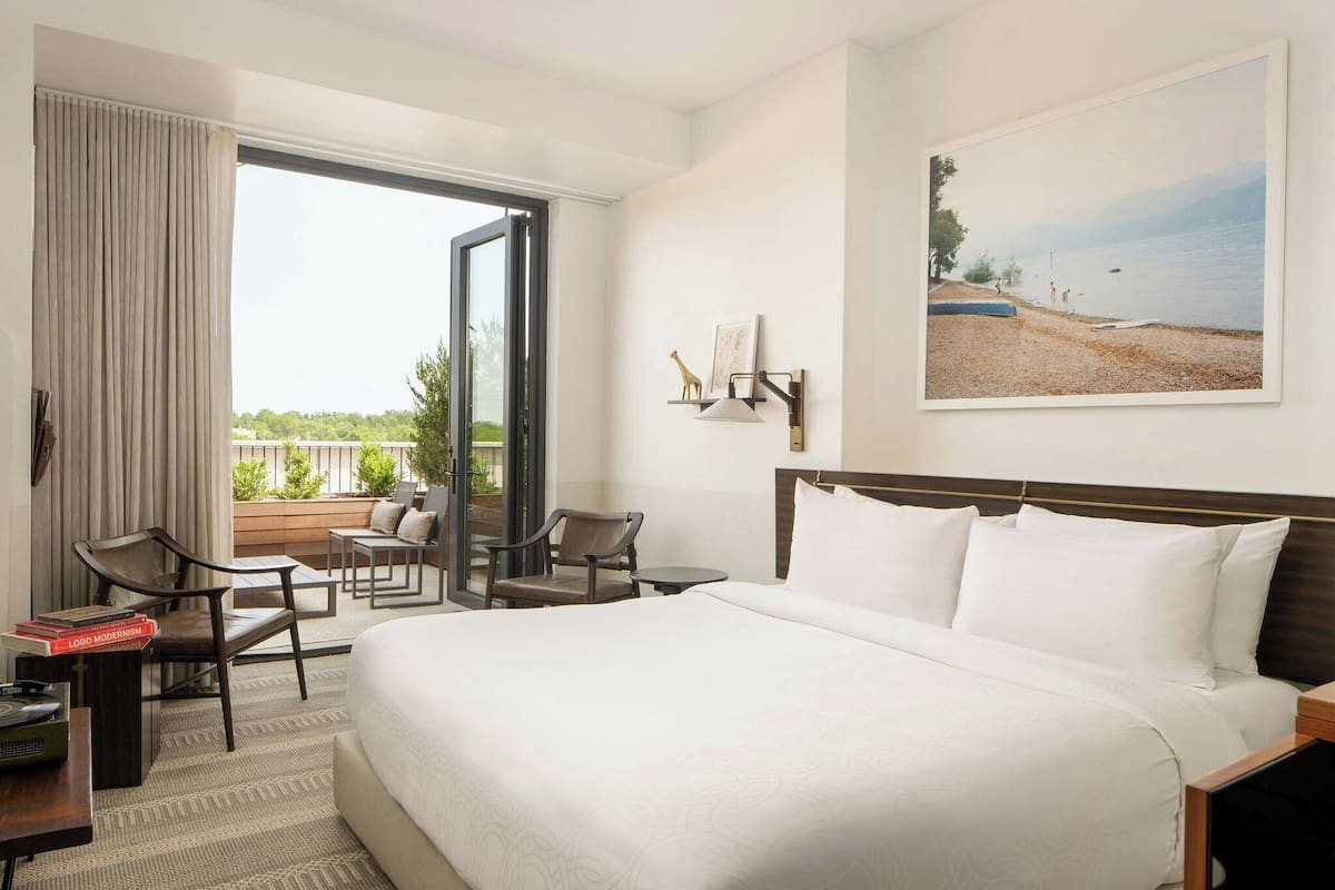 A hotel room with a large bed, a desk with a chair, a small table, and a balcony with chairs.