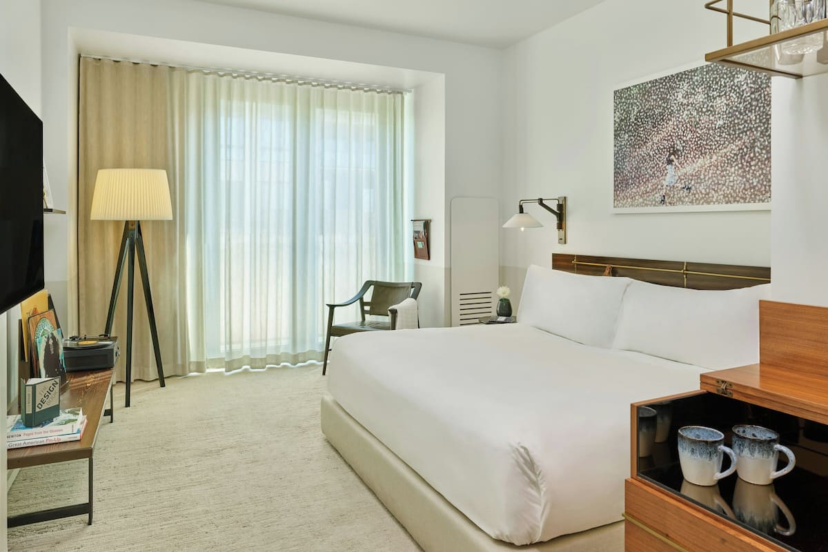 A hotel room with a large bed, a desk with a lamp, a chair, a bookshelf, a painting on the wall, and a window with curtains.