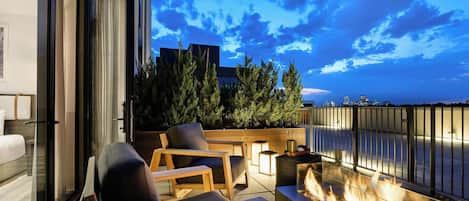 A balcony with a fire feature, seating, and a view of the cityscape at dusk.