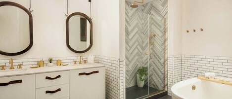 Un baño moderno con tocador blanco, detalles dorados, un espejo grande y una ducha con diseño de espiga.