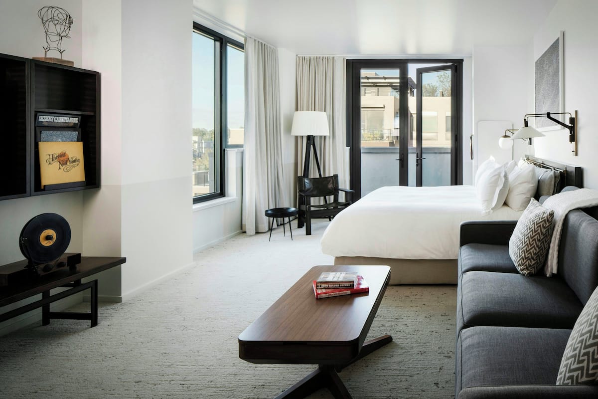 A hotel room with a bed, a desk, a chair, a lamp, and a view of the outside.