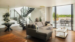 A modern living room with a sectional sofa, a glass coffee table, a wooden bench, a staircase with glass railings, and a view of the outdoors. - Halcyon, a hotel in Cherry Creek, Denver (Denver)
