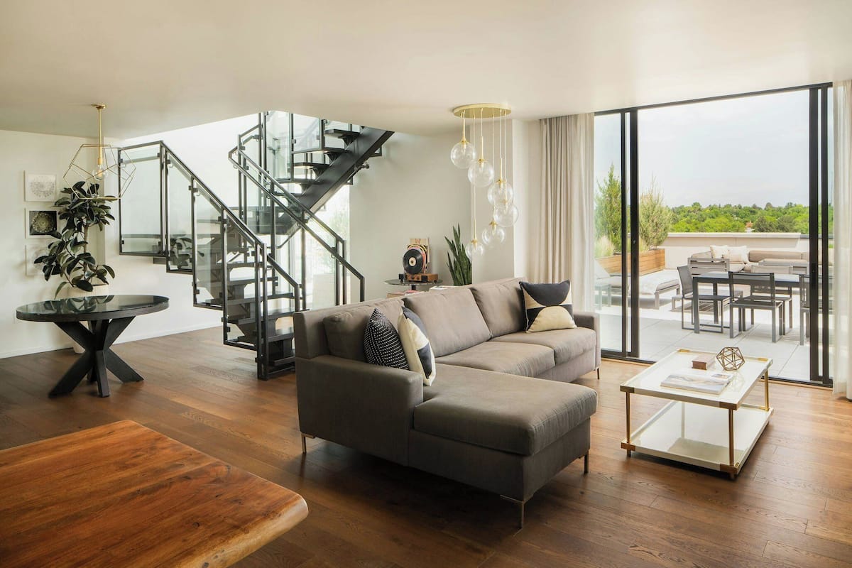 A modern living room with a sectional sofa, a glass coffee table, a wooden bench, a staircase with glass railings, and a view of the outdoors.