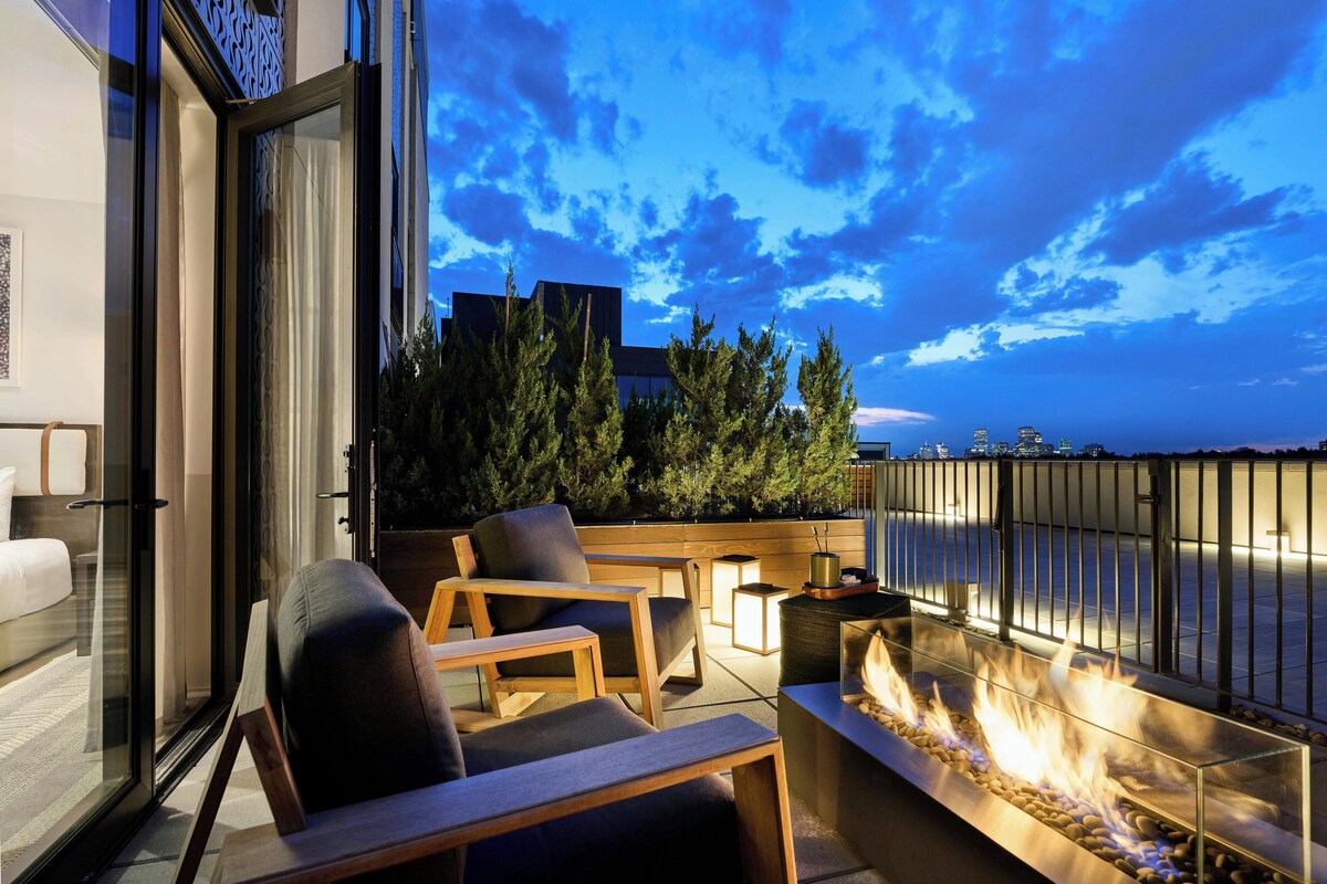 Balcón con una chimenea exterior, asientos y vistas del horizonte urbano al anochecer.