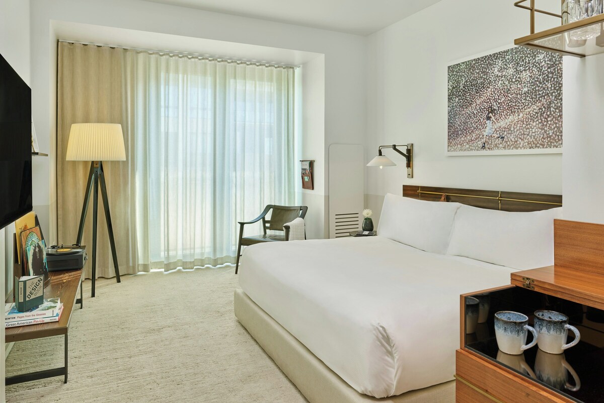 A hotel room with a bed, a desk with a lamp, a chair, a coffee table with mugs, and a painting on the wall.