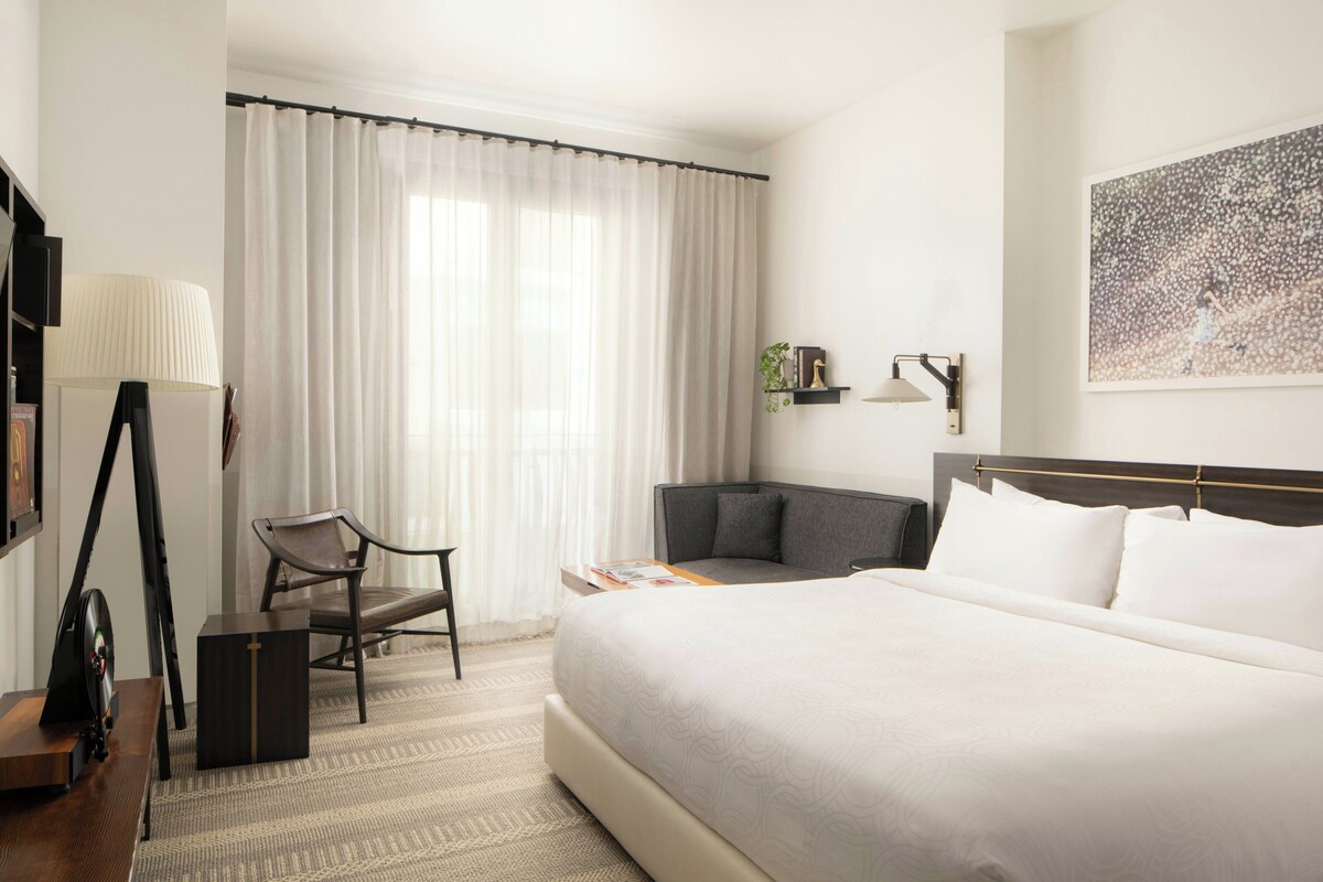 A hotel room with a large bed, a desk, a chair, a lamp, and a painting on the wall.