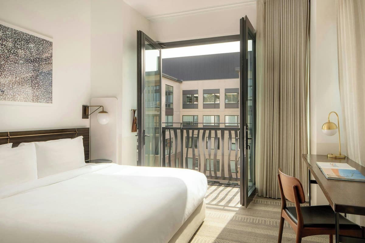 A hotel room with a bed, a desk, a chair, a window with a view of buildings, and a door leading to an outdoor balcony.