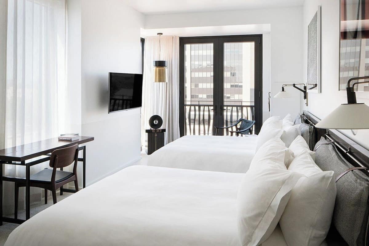 A hotel room with two beds, a desk, a chair, a TV, a balcony, and a lamp.